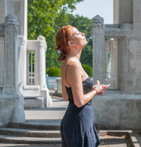 A white woman with red hair turns her face to the sun, eyes closed. Her hands are holding a cellphone in front of her, and there are stone columns in the background.